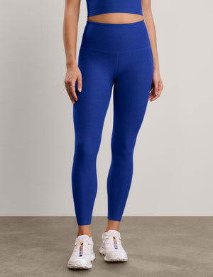 A woman modeling a matching royal blue activewear set consisting of a cropped tank and high-waisted leggings, paired with white sneakers, against a neutral studio backdrop.