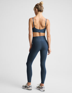 A woman shown from the back wearing a matching navy blue activewear set, consisting of a sports bra and high-waisted leggings, paired with white and gray sneakers, against a light gray studio background.