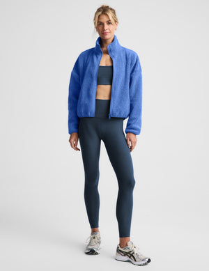 A woman modeling a bright blue cropped fleece zip-up jacket over a dark gray sports bra and matching dark gray leggings, paired with white and gray running shoes, against a light gray studio background.