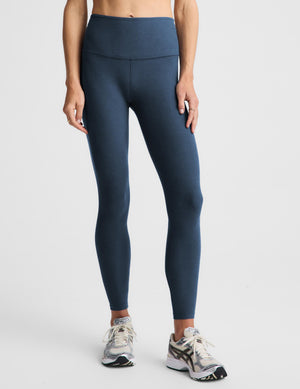 A model wearing high-waisted, full-length athletic leggings in a dark slate blue color, paired with gray and white running shoes, against a light gray studio background.