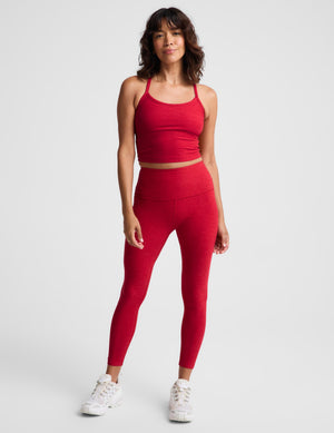 A woman modeling a matching red activewear set consisting of a crop top and high-waisted leggings, paired with white sneakers, against a light gray studio background.