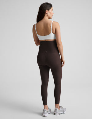 Pregnant woman wearing a white bra and brown maternity midi leggings against a plain studio background. 