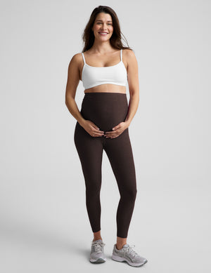 Pregnant woman wearing a white bra and brown maternity midi leggings against a plain studio background. 