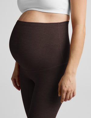 Pregnant woman wearing a white bra and brown maternity midi leggings against a plain studio background. 