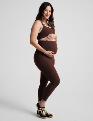 Pregnant woman wearing a brown sports bra and brown maternity leggings against a plain background. 