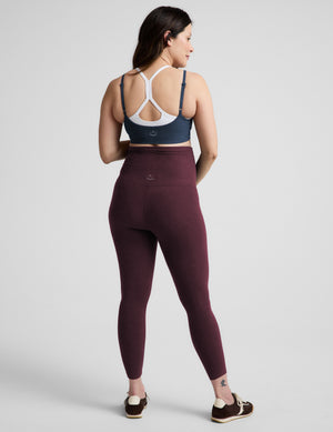 A pregnant model wearing high-waisted burgundy maternity leggings, a navy blue sports bra, and black sneakers with white laces, standing against a light gray studio background.