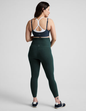 A woman modeling a dark green high-waisted leggings set with a blue sports bra, standing against a light gray studio background.