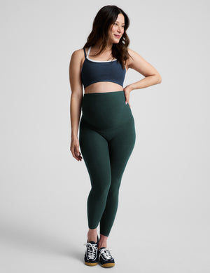 A woman modeling a dark green high-waisted leggings set with a blue sports bra, standing against a light gray studio background.