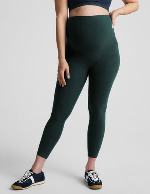 A woman modeling a dark green high-waisted leggings set with a blue sports bra, standing against a light gray studio background.