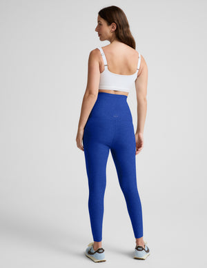 A woman is shown from the back wearing high-waisted royal blue leggings and a white sports bra, standing against a light gray studio background.