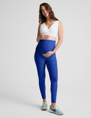 A pregnant woman posing in high-waisted royal blue maternity leggings, a white wrap-style crop top, and light blue sneakers against a light gray studio background.