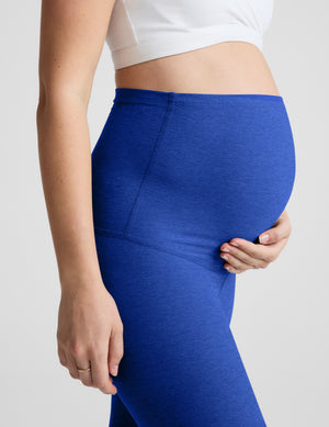 A close-up of a pregnant model wearing high-waisted royal blue maternity leggings and a white sports bra against a light gray studio background.