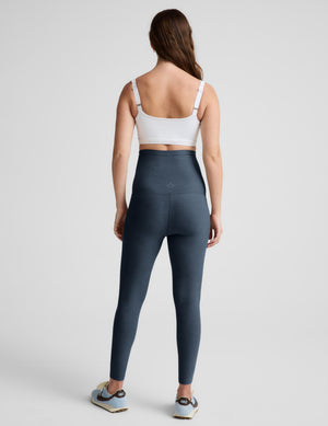 A pregnant woman modeling high-waisted, full-length leggings in a slate blue-gray color, paired with a white wrap-style sports bra and light blue sneakers, against a solid light gray studio background.