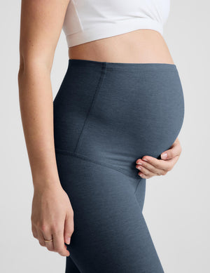 A pregnant woman modeling high-waisted, full-length leggings in a slate blue-gray color, paired with a white wrap-style sports bra and light blue sneakers, against a solid light gray studio background.