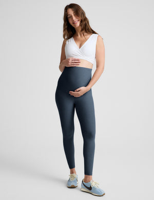 A pregnant woman modeling high-waisted, full-length leggings in a slate blue-gray color, paired with a white wrap-style sports bra and light blue sneakers, against a solid light gray studio background.