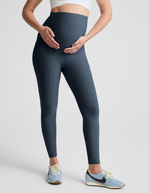 A pregnant woman modeling high-waisted, full-length leggings in a slate blue-gray color, paired with a white wrap-style sports bra and light blue sneakers, against a solid light gray studio background.