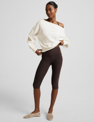 Woman wearing a brown bra, pedal pusher leggings, and a white off the shoulder fleece sweatshirt against a plain background. 