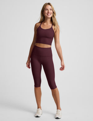 A woman modeling a matching burgundy activewear set consisting of a cropped tank and high-waisted capri leggings, paired with white sneakers, against a light gray studio background.