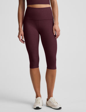 A model wearing high-waisted, heathered burgundy capri leggings and cream athletic sneakers, shown from the waist down against a light gray studio background.