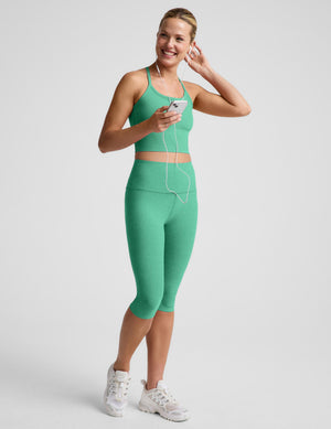 A woman modeling a matching green activewear set consisting of a cropped tank and high-waisted capri leggings, paired with white sneakers, against a light gray studio background.