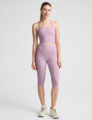 A woman modeling a matching dusty purple activewear set consisting of a cropped tank top and high-waisted capri leggings, paired with beige sneakers, against a light gray studio background.