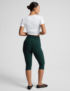 A woman modeling dark green high-waisted capri leggings with a white t-shirt and black flat shoes, standing against a light gray studio background.