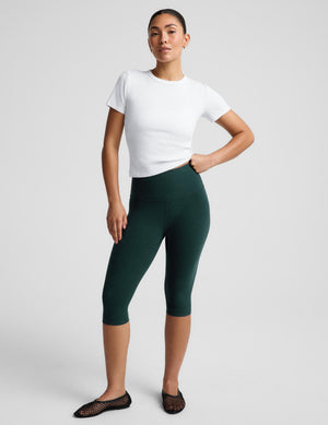 A woman modeling dark green high-waisted capri leggings with a white t-shirt and black flat shoes, standing against a light gray studio background.