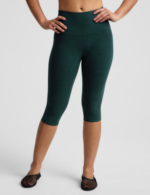 A model wearing dark green high-waisted capri leggings and black mesh flat shoes, standing against a light gray studio background.