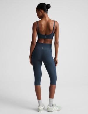 A woman shown from the back wearing a matching dark slate blue activewear set consisting of a sports bra and high-waisted capri leggings, paired with white socks and light green sneakers, against a light gray studio background.