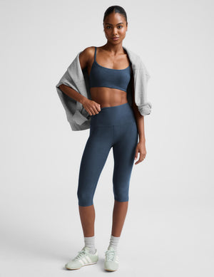 A woman modeling a matching slate blue activewear set consisting of a sports bra and high-waisted capri leggings, with a light gray cardigan draped over her shoulders, white socks, and white sneakers against a light gray studio background.