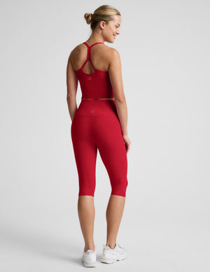 A woman wearing a matching red activewear set consisting of a sports bra and high-waisted capri leggings, shown from a rear three-quarter angle against a light gray studio background.