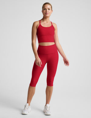 A woman modeling a matching red activewear set consisting of a cropped tank and high-waisted capri leggings, paired with white sneakers, against a light gray studio background.