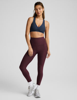 A woman modeling a matching activewear set consisting of a navy blue sports bra and high-waisted burgundy leggings, paired with white socks and gray running shoes, against a light gray studio background.