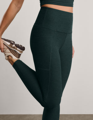 A close-up of a model wearing dark forest green high-waisted leggings, stretching her leg while holding a New Balance sneaker against a neutral studio background.