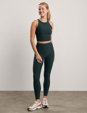 A woman modeling a matching dark green athletic set consisting of a sleeveless crop top and high-waisted leggings, paired with white and pink sneakers, against a neutral studio backdrop.