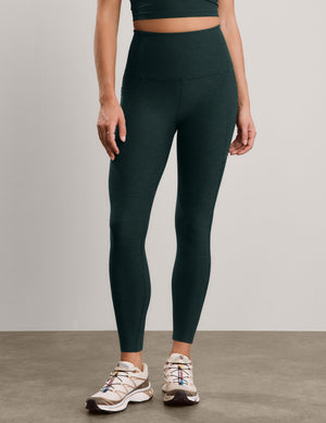 A model wearing dark forest green high-waisted leggings and beige athletic sneakers, standing against a neutral light gray studio background.