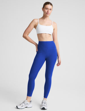 A woman modeling high-waisted royal blue leggings with a white sports bra and white sneakers, standing against a light gray studio background.