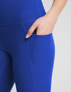 A woman modeling high-waisted royal blue leggings with a white sports bra and white sneakers, standing against a light gray studio background.