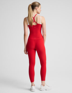 A woman modeling a matching red activewear set consisting of a sleeveless halter crop top and high-waisted leggings, paired with white sneakers, against a light gray studio background.