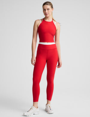 A woman modeling a matching red activewear set consisting of a sleeveless halter crop top and high-waisted leggings, paired with white sneakers, against a light gray studio background.