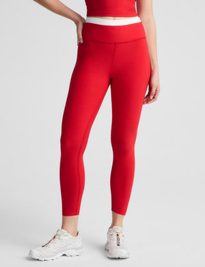 A woman modeling a matching red activewear set consisting of a sleeveless halter crop top and high-waisted leggings, paired with white sneakers, against a light gray studio background.