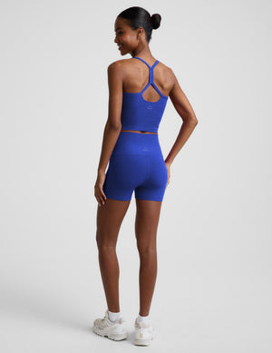 A woman modeling a matching athletic set consisting of a royal blue tank top and high-waisted shorts, paired with white sneakers, against a light gray studio background.