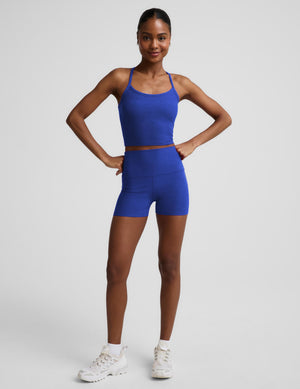 A woman modeling a matching athletic set consisting of a royal blue tank top and high-waisted shorts, paired with white sneakers, against a light gray studio background.