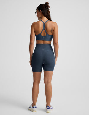 A woman is shown from the back wearing a matching athletic set consisting of a dark slate blue sports bra and high-waisted biker shorts, paired with white sneakers, against a light gray studio background.