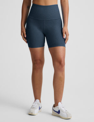 A model from the waist down wearing high-waisted, fitted athletic biker shorts in a heathered slate blue color, paired with white and blue sneakers, against a light gray studio background.