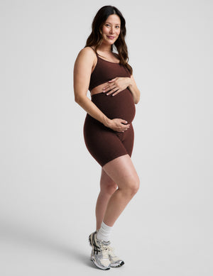 Pregnant woman wearing a brown sports bra and brown maternity biker shorts against a plain background. 