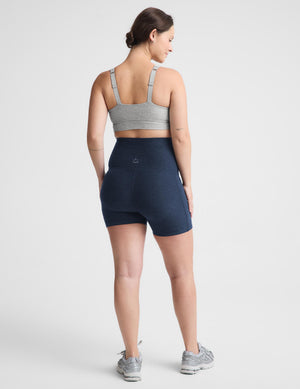 Pregnant woman wearing a gray sports bra and navy biker shorts with pockets against a plain background. 
