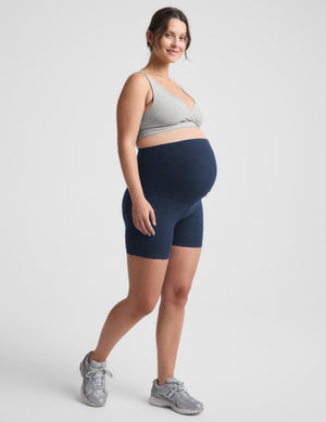 Pregnant woman wearing a gray sports bra and navy biker shorts with pockets against a plain background. 