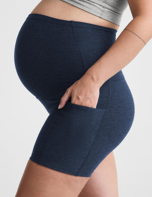 Pregnant woman wearing a gray sports bra and navy biker shorts with pockets against a plain background. 