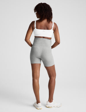 Pregnant woman wearing a white maternity bra and gray biker shorts with pockets against a plain background. 